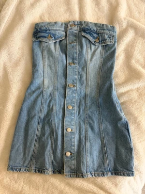 Jeansklänning - Säljer en snygg blå jeansklänning i kort modell med knappar hela vägen framtill och fickdetaljer upptill. Klänning har dragkedja i sidan och är gjord i klassisk denim. Perfekt till sommaren!🥰