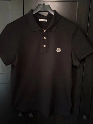 Svart Moncler pikétröja i bomull - Säljer en svart pikétröja från Moncler med klassisk krage och tre knappar framtill. Tröjan har Monclers ikoniska logga broderad på bröstet och är gjord i mjuk bomull. Perfekt för en clean och stilren look.