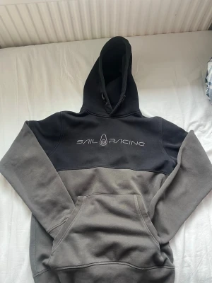 Grå och svart hoodie från Sail Racing - Säljer en snygg tvåfärgad hoodie från Sail Racing i storlek S. Hoodien är svart upptill och grå nedtill, har stor magficka och justerbar huva. Loggan är broderad på bröstet. Perfekt för dig som gillar sportig och avslappnad stil.