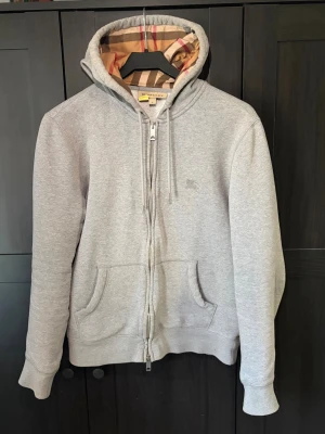 Grå zip hoodie från Burberry - Säljer en grå hoodie från Burberry med dragkedja och klassiskt rutigt foder i huvan. Hoodien har två stora fickor fram, justerbar dragsko i huvan och diskret Burberry-logga på bröstet. Tillverkad i mjuk bomullsblandning för extra komfort.