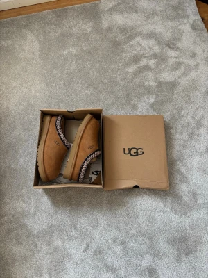 Bruna UGG tasman i mocka - Säljer ett par bruna UGG tofflor i mjuk mocka med broderad kant i svart, vitt och beige. Tofflorna har en klassisk slip-on modell och en rejäl sula som passar perfekt för att chilla hemma. UGG-logga på sidan. Kommer i originalkartong.