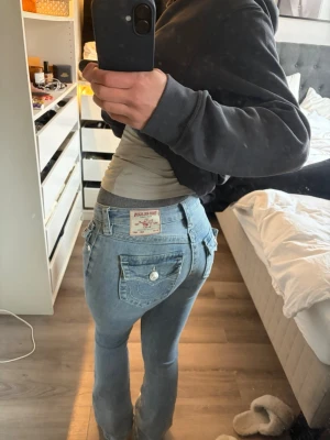 Ljusblå True Religion jeans bootcut - Säljer ett par ljusblå jeans från True Religion med bootcut-modell och klassiska kontrastsömmar. Jeansen har dekorativa ficklock bak med knappar och tydlig logga på bakfickan. Tillverkade i denim med en snygg tvätt och låg midja.