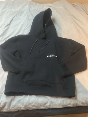 Svart hoodie från Sail Racing - Svart hoodie från Sail Racing med klassisk huva och dragsnören. Framtill finns en stor magficka och en liten logga på bröstet. Tillverkad i mjukt material som känns skönt mot huden. Perfekt för en avslappnad och stilren look.