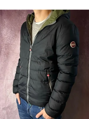 Svart pufferjacka från Colmar - Snygg svart pufferjacka från Colmar med huva och dragkedja framtill. Jackan har två fickor med dragkedja och en diskret logga på ärmen. Insidan av huvan är olivgrön för en cool kontrast. Perfekt för kalla vårdagar.