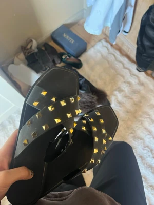 Svarta sandaler med guldfärgade nitar - Coola svarta sandaler  med två breda remmar dekorerade med guldfärgade pyramidnitar. Sandalerna är tillverkade i syntetmaterial och har en platt sula som är bekväm att gå i. 