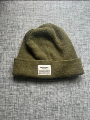 Olivgrön mössa från Resteröds - Beanie / mössa från Resteröds med uppvikt kant och vit tygpatch framtill. Onesize. Material: Akryl