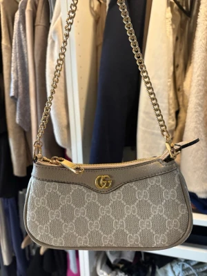 Beige axelremsväska - Snygg beige handväska med mönster i canvas och detaljer i läder. Väskan har guldfärgad kedjerem och dragkedja upptill. Aldrig använd 