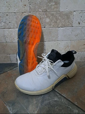 Golfskor från Ecco - Snygga vita Golfskor med svart häl och färgglad sula i orange och blått. Skorna har lågt skaft, vita snören och en robust yttersula med bra grepp. Perfekta för dig som vill sticka ut med coola detaljer och sportig look.