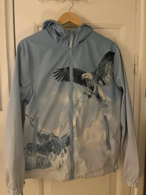 Trapstar Eagle Vindjacka - Sky Blue Eagle vindjacka från Trapstar. Jackan har huva med snörning och dragkedja framtill. Perfekt för dig som vill sticka ut med en unik design och sportig look. Endast använd ett fåtal gånger. Om du är storlek S är M den perfekta storleken för dig när det kommer till denna jackan.