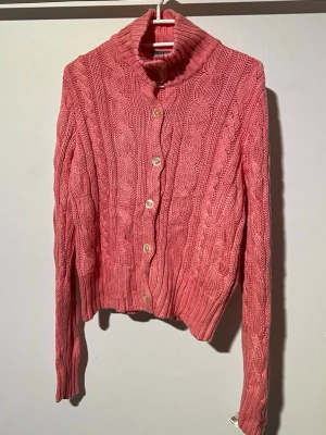 Rosa kabelstickad kofta med knappar - Säljer en rosa kabelstickad kofta med stora vita knappar och hög krage. Koftan har långa ärmar och ribbade muddar vid ärmslut och nederkant. Perfekt för dig som gillar mysiga och trendiga plagg med klassisk känsla.