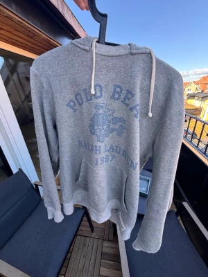 Grå hoodie Polo Bear Ralph Lauren🙌🤩 - Snygg grå hoodie från Ralph Lauren med Polo Bear-tryck och texten '1967' framtill. Hoodien har huva med vita dragsnören och en stor känguruficka. Tillverkad i mjukt sweatshirtmaterial, perfekt för en avslappnad streetwear-look.🙌🤩