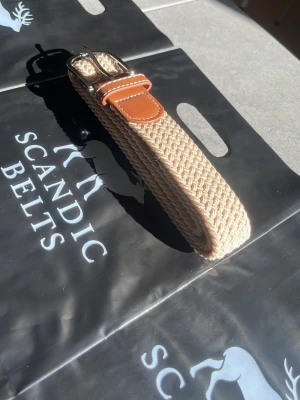 Beige flätat bälte  - Snyggt beige flätat bälte med detaljer i brunt skinn och silverfärgat spänne. Perfekt accessoar för att lyfta din outfit med en stilren och modern touch. Det är 105 cm långt.