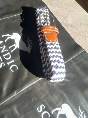 Randigt flätat bälte med läderdetalj - Snyggt flätat bälte i vitt och marinblått med randigt mönster. Bältet har en silverfärgad metallspänne och bruna läderdetaljer vid spännet. Perfekt accessoar för att lyfta din outfit. 105cm