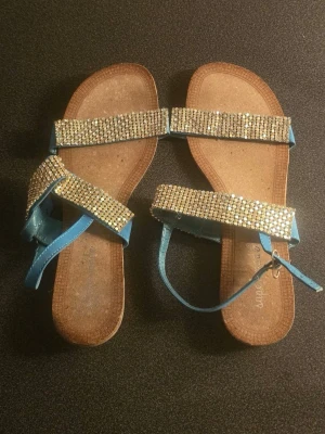 Blå sandaler med strassdetaljer - Snygga sandaler i blått med breda remmar täckta av glittrande strass. Remmarna är i konstläder och sandalerna har en öppen tå-design samt justerbart spänne vid ankeln. Perfekta för dig som vill sticka ut med lite extra bling.