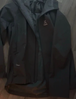 grå regnjacka från Haglöfs - Snygg grå regnjacka från Haglöfs med huva och dragkedja framtill. Jackan har diskret logga på bröstet och är tillverkad i ett vattentätt material, perfekt för regniga dagar. Innerfodret har ett mönster och jackan har praktiska fickor.