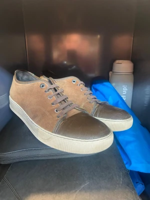 Lanvins  - Snygga bruna sneakers med lågt skaft och mockaliknande material på ovandelen. Tåpartiet och snörningen är i mörkare brunt läderimitation. Vita, slitstarka gummisulor och klassisk snörning med metallöljetter. Perfekta för en avslappnad streetstil.