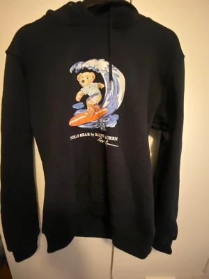 Svart hoodie Polo Bear Ralph Lauren (ej äkta) - Svart hoodie från Ralph Lauren med Polo Bear-tryck där björnen surfar på en våg. Klassisk huva med dragsko och ribbade muddar. Mjuk bomullskänsla och avslappnad passform. Perfekt för dig som gillar streetwear och snygga prints. Hoodien är lite oversized så den funkar också som M