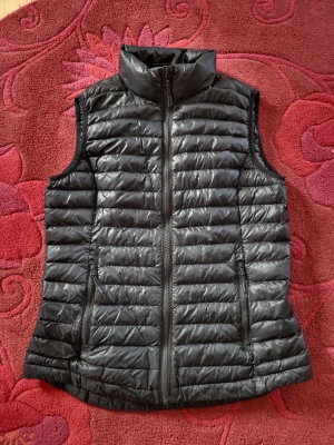 Svart lättviktsväst med dragkedja - Snygg svart dunväst i lättviktsmodell med horisontella sömmar och hög krage. Västen har två dragkedjeförsedda fickor framtill och stängs med en hel dragkedja. Perfekt för lager-på-lager och enkel att matcha till olika outfits.