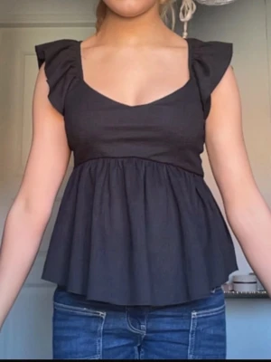 Svart volangärmad topp - Snygg svart topp med volangärmar och v-ringning. Toppen har en lös passform nedtill och markerad midja som ger en feminin siluett. Perfekt att styla med jeans eller kjol för en trendig look. Aldrig använd 
