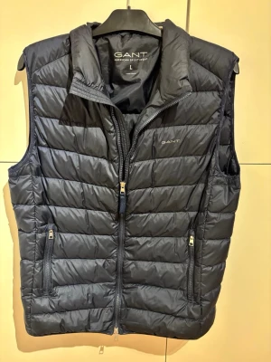 Mörkblå dunväst från GANT - Snygg mörkblå dunväst från GANT med quiltad design och hög krage. Västen har två dragkedjeförsedda fickor fram och diskret GANT-logga på bröstet. Tillverkad i lätt och värmande material, perfekt för lager på lager under kyliga dagar. Köpt för 1499, mitt pris 699🤗