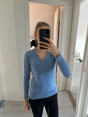 Ljusblå v-ringad tröja Tommy Hilfiger - Snygg ljusblå långärmad tröja från Tommy Hilfiger med v-ringning och diskret logga på bröstet. Tröjan har en smal passform.