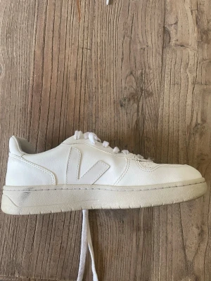 Vita Veja sneakers i läder - Säljer ett par klassiska vita Veja sneakers i läder med perforerad tå och tydlig V-logga på sidan. Skorna har vita skosnören och en vit sula som ger en clean och stilren look. Perfekta för dig som gillar minimalistisk och trendig stil.