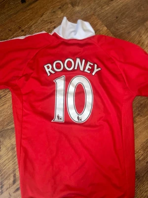 Manchester United Rooney tröja Nike retro - Röd Manchester United matchtröja från Nike med tryck 'ROONEY 10' på ryggen och AON som sponsor på framsidan. Klassisk vit krage och klubbmärke på bröstet. Perfekt för fotbollsfans som vill visa sitt stöd för laget. Äkta autentic