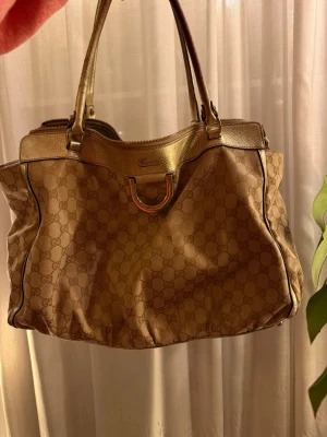 Beige handväska från Gucci - Säljer en beige handväska från Gucci med klassiskt GG-mönster och detaljer i guldigt läder. Väskan har dubbla handtag, metallspänne framtill och rymlig form. Perfekt för dig som gillar lyxiga accessoarer och tidlös design.