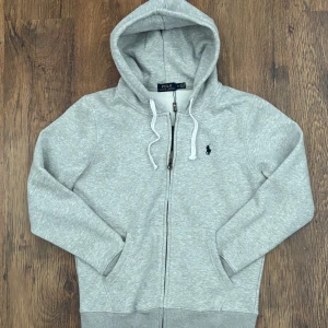 Grå zip Hoodie från Polo Ralph Lauren - Snygg grå zip-hoodie från Polo Ralph Lauren med huva och vita dragsnören. Klassisk design med broderad logga på bröstet och mjukt material som känns skönt mot huden. Perfekt för en avslappnad och stilren look.