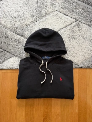 Svart hoodie från Polo Ralph Lauren - Svart hoodie från Polo Ralph Lauren med klassisk huva och vita dragsnören. På bröstet finns den ikoniska röda Polo-loggan broderad. Hoodien har en stor magficka och ribbade muddar vid ärmslut och nederkant. Perfekt för en avslappnad och stilren look.