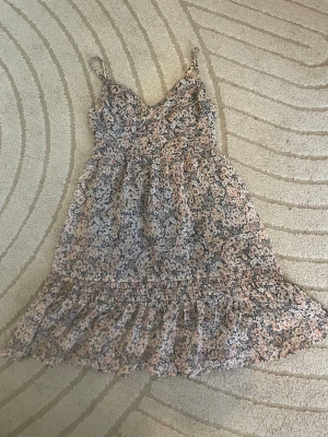 Blommig mesh hollister sommaklänning❤️ - Söt kort klänning med smala axelband och volangkant nertill. Klänningen har ett blommigt mönster i ljusa toner av beige, vit och svart. Perfekt för varma dagar och har en luftig passform. Materialet  känns lätt och somrigt.