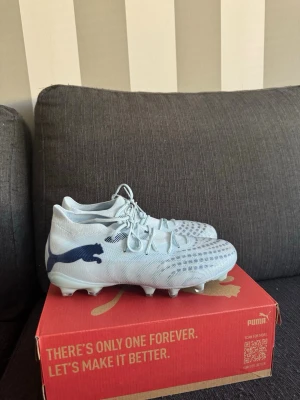 Vita Puma fotbollsskor med blå logga - Snygga ijus blå fotbollsskor från Puma med blå logga på sidan. Skorna har snörning, låg profil och mönstrad ovandel för bättre bollkontroll. Tillverkade i syntetmaterial och designade för spel på gräs. Perfekta för dig som vill sticka ut på planen.