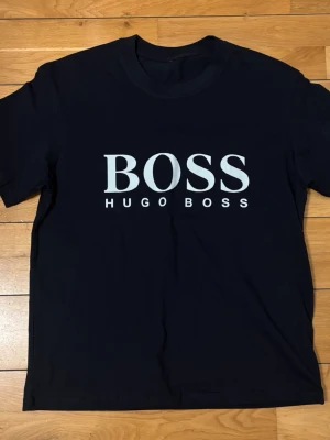 Svart Hugo Boss t-shirt med logga - Snygg svart t-shirt från Hugo Boss med stor vit BOSS-logga och texten HUGO BOSS på bröstet. Klassisk rund halsringning och rak passform. Tillverkad i mjukt bomullsmaterial som känns skönt mot huden. Perfekt för en stilren och enkel look.