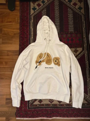 Vit Palm Angels hoodie med björntryck - Säljer en vit hoodie från Palm Angels med ett unikt björntryck på bröstet och svart text. Hoodien har stor huva med dragsko och en klassisk magficka. Tillverkad i mjukt bomullsmaterial och har ribbade muddar vid ärmslut och nederkant. Perfekt för en chill och trendig look.
