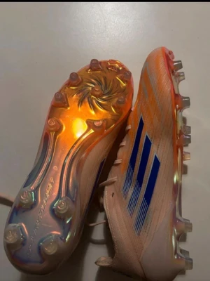 Adidas F50 Sparkfusion fotbollsskor - Säljer ett par Adidas F50 fotbollsskor med Speedframe-sula. Skorna är ljusorange med blå ränder och detaljer, samt har en skimrande, iriserande yttersula. Lätta och smidiga med snörning, perfekta för snabba spelare på planen. De är helt nya och aldrig använts. 