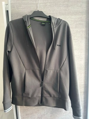 Mörkgrå hoodie från BOSS - Snygg mörkgrå hoodie från BOSS med dragkedja framtill och två stora fickor. Klassisk huva med dragsko och diskret BOSS-logga på bröstet. Detaljer i grönt på insidan av kragen ger en cool kontrast. Perfekt för en avslappnad och stilren look.