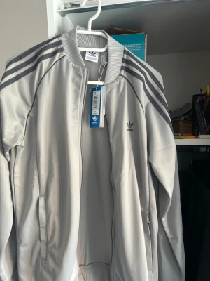 Grå adidas kofta - Sportig ljusgrå kofta från adidas med klassiska mörkgrå ränder längs ärmarna och adidas-logga på bröstet. Koftan har dragkedja framtill, ribbade muddar och är tillverkad i polyester för en bekväm och stilren look.