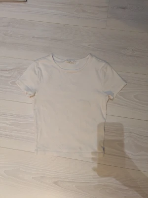 Vit basic t-shirt från Basically Basics/ Gina Tricot  - En enkel och stilren vit t-shirt från Basically Basics/Gina Tricot i storlek XS. T-shirten har rund halsringning och korta ärmar, perfekt till alla outfits. Tillverkad i mjuk bomull för en skön känsla hela dagen. T-shirten är i kortare slag och kan därför vara som en magtröja beroende på kroppsform. 44 cm lång.