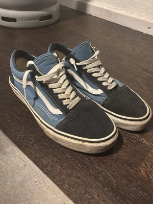Blå och svarta Vans Old Skool sneakers - Säljer ett par klassiska Vans Old Skool sneakers i blått och svart med vit sula och vita snören. Skorna har den ikoniska vita sidan, tåparti i mocka och canvas på sidorna. Perfekta för dig som gillar streetwear och vill ha en tidlös look.