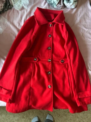 Röd ullkappa med stora knappar - Stilren röd ullkappa med markerad midja och stora, silvriga knappar framtill. Kappan har klassisk krage och dekorativa knappar vid midjan för extra detalj. Perfekt för kyliga dagar när du vill sticka ut. 