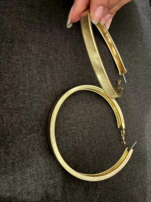 Stora guldiga creoler örhängen - Säljer ett par stora, runda creoler i guldfärgad metall. Örhängena har en bred, räfflad yta som ger en snygg och modern look. Perfekta för dig som vill ha ett statement-smycke till din outfit. Lätta att bära och stängs med klassiskt nål-lås.