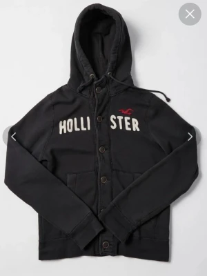 Svart hoodie från Hollister med knappar - Mörkgrå hoodie från Hollister med stora vita bokstäver och röd logga på bröstet. Tröjan har huva, knäppning med träknappar och två fickor framtill. Tillverkad i mjukt bomullsmaterial som känns riktigt skönt mot huden. Xl i storlek, men passar nu L