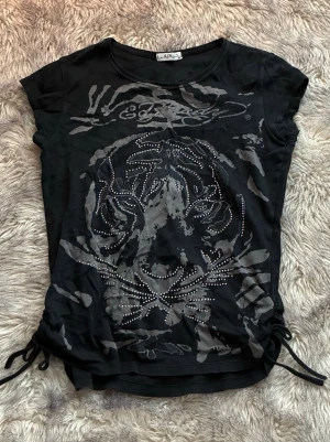 Unik Ed Hardy t-shirt - Supersnygg svart t-shirt från Ed Hardy med stort tigertryck framtill dekorerat med silvriga nitar. Storlek XL men den sitter som S-M! Tröjan har korta ärmar, snörning vid sidorna och Ed Hardy på ryggen. Snörningen går att justera 🖤