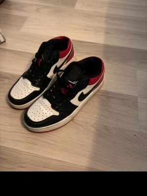 Nike Air Jordan 1 Low svart/vit/röd - Säljer ett par Nike Air Jordan 1 Low sneakers i svart, vitt och rött. Skorna har klassisk låg siluett, svart swoosh, perforerad tåbox och röda detaljer runt hälen. Tillverkade i läder med snörning framtill och vit sula med röd kant.