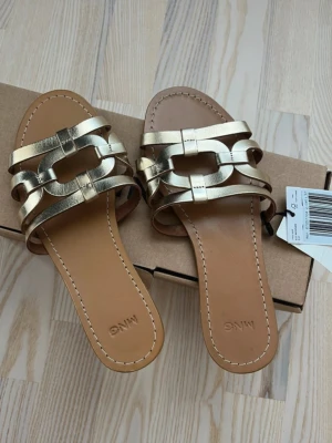 Guldiga lädersandaler från Mango storlek 37 - Snygga slip-in sandaler från Mango i metallic guld med breda remmar och dekorativa länkar. Sandalerna har öppen tå och platt sula i brunt konstläder. Perfekta för en trendig och avslappnad look under sommaren.