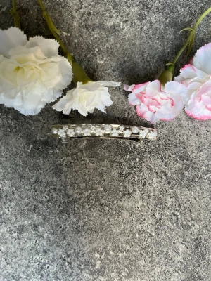 Handgjort hårspänne med pärlor - Säljer ett elegant hårspänne i silverfärg dekorerat med små vita blompärlor och glittrande stenar. Perfekt accessoar för att ge frisyren en extra touch av stil. Enkelt spänne att fästa i frisyren 🥰✨ dessa hårspännens design är handgjorda och har satts på sten bit för bit. 