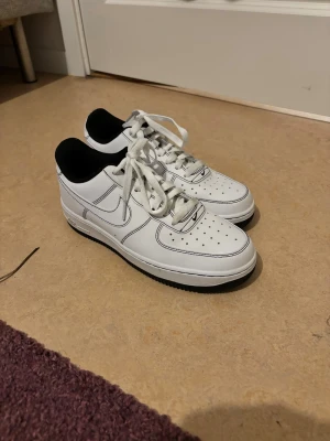 Nike Air Force 1 vita sneakers - Klassiska vita Nike Air Force 1 sneakers med svart detalj på insidan och svart sula. Skorna har perforerad tå, vita skosnören och tydliga sömmar. Helt nya använt 2 gånger 