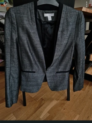 Grå kavaj med svart krage - Snygg grå kavaj från H&M med svart kontrastkrage och smal passform. Kavajen har två dekorativa fickor framtill och är helfodrad med svart innerfoder. Perfekt för en stilren och modern look.