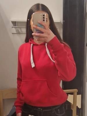 Röd/rosa hoodie med vita snören - Säljer en klassisk röd hoodie med vita dragsnören och stor känguruficka framtill. Hoodien har en avslappnad passform och ribbade muddar vid ärmslut och nederkant. Perfekt för en chill och stilren look.