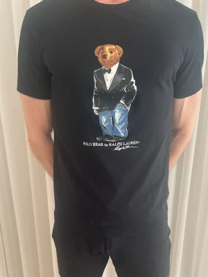 Svart Polo Bear t-shirt från Ralph Lauren - Svart t-shirt från Ralph Lauren med ikoniska Polo Bear-trycket på bröstet. Björnen har på sig smoking och jeans, vilket ger en cool kontrast. T-shirten har rund hals och korta ärmar. Perfekt för dig som gillar streetstyle och klassiska märken.